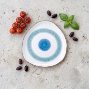 Top-down view of an Aegean Eye ceramic plate surrounded by cherry tomatoes, olives, and fresh basil on a stone surface.